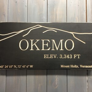 May include: A rectangular wooden sign with a black background and white engraved text and mountain outline. The sign reads "OKEMO ELEV. 3,343 FT" and includes coordinates and "Mount Holly, Vermont".