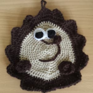 Puede incluir: Un agarrador de olla hecho a mano, tejido a ganchillo con forma de león sonriente. El león tiene una melena marrón, una cara beige y ojos negros y blancos. El agarrador tiene un lazo para colgar.