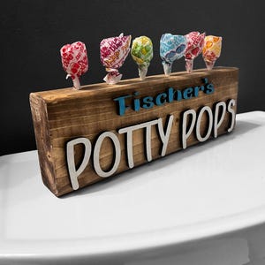 May include: A rectangular wooden block with a dark stained finish featuring the text "Fischer's" in blue and "POTTY POPS" in white raised lettering. Six colorful lollipops are inserted into holes along the top edge of the wooden block.