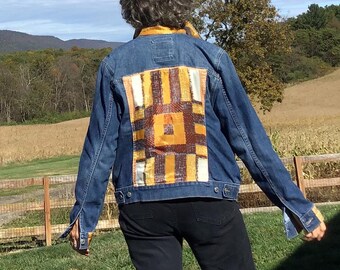 May include: A denim jacket with a colorful patchwork design on the back. The jacket is blue with a square pattern of brown, yellow, and white fabric. The person is standing in a field with a blue sky and trees in the background.
