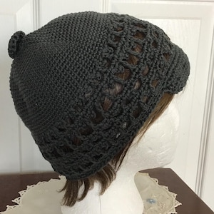 May include: Dark gray crocheted hat with a textured design and a small pom-pom on top. The hat features an openwork pattern around the lower edge. The hat is displayed on a white mannequin head.