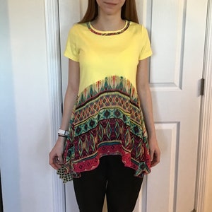 May include: A yellow short-sleeved shirt with a colorful patterned bottom hem. The shirt has a high, round neckline.