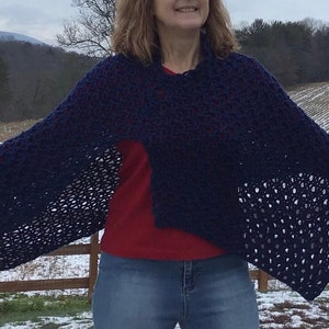 May include: A woman wearing a navy blue crochet poncho with a red shirt and blue jeans. She is standing in a snowy field with a wooden fence in the background.