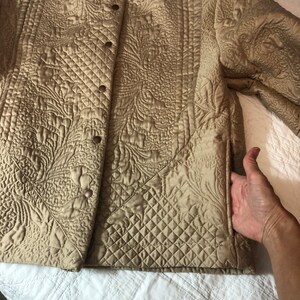 May include: A quilted jacket in a light brown color with a floral pattern. The jacket has a button closure and a pocket on the right side.