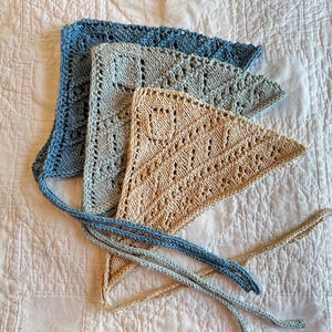 May include: Three triangular headscarves made of knitted fabric with a lace-like pattern. The scarves are layered and colored in shades of dark blue, light blue, and beige. Each scarf features long knitted ties extending from the bottom corners.