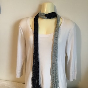 May include: A black and gray knitted scarf with fringe on the ends. The scarf is tied around a white mannequin's neck.
