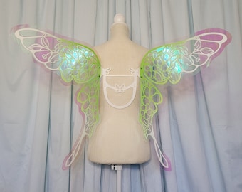 Puede incluir: Un par de alas de hada iridiscentes con un diseño floral verde y blanco. Las alas están unidas a un marco de plástico blanco.