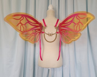 Puede incluir: Un par de alas de hada iridiscentes con un degradado de color dorado y rosa. Las alas están unidas a un arnés de metal dorado con correas de cinta rosa.