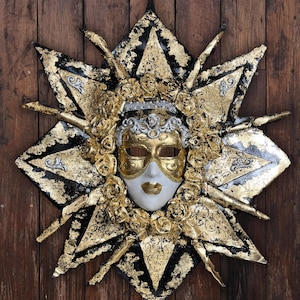 Pode incluir: Uma máscara veneziana decorativa com detalhes dourados, prateados e pretos, colocada sobre um desenho em forma de estrela. A máscara apresenta um rosto branco, olhos dourados e um lábio dourado. A máscara é rodeada por enfeites florais dourados.