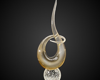 May include: A clear glass sculpture with a teardrop shape and a curved, pointed top. The sculpture sits atop a clear, bubble-filled sphere. The glass has a golden hue, and the background is black.