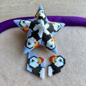 May include: A set of three beaded ornaments in the shape of a star and two diamonds. The ornaments are blue, white, black, orange, and yellow and feature a pattern of puffins.