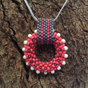 Może przedstawiać: Naszyjnik z koralików z okrągłym, czerwono-białym wisiorem. Wisiorek wykonany jest z małych koralików i ma szarą i różową wstążkę z koralików.