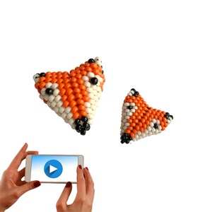 May include: Two orange, white, and black beaded fox shapes. The larger fox is in the shape of a heart. The smaller fox is in the shape of a triangle. A hand is holding a smartphone with a play button on the screen.