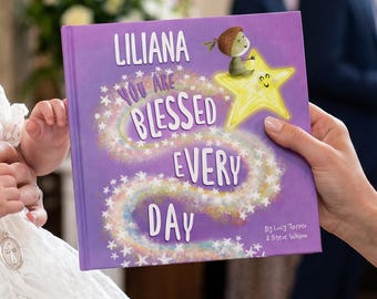 May include: A purple children's book titled "Liliana You Are Blessed Every Day." The cover features a cartoon illustration of a child on a star, with a swirling design and text in white. The book is held by a person.