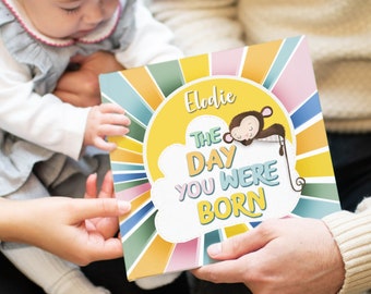 Könnte beinhalten: Ein personalisiertes Buch in Gelb, Blau, Grün und Rosa mit dem Text "The Day You Were Born" und einer Illustration eines schlafenden Affen. Das Buch trägt den Titel "Elodie".