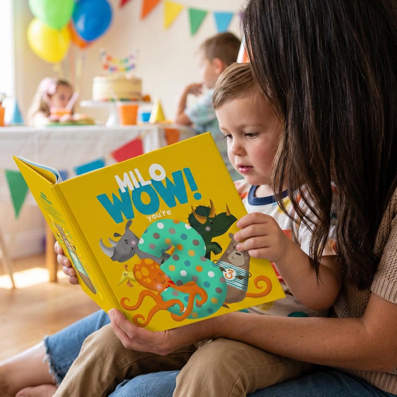 May include: A yellow children's book titled "Milo Wow! You're 3" is being read by a child. The book features colorful illustrations of animals and a large number 3. The scene is set at a birthday party with balloons and a cake in the background.