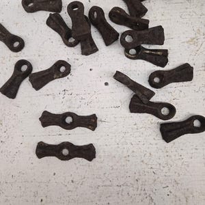 May include: A selection of small, antique, rusty brown metal hardware pieces. Each piece features a loop at one end and a decorative shape at the other. The hardware is scattered on a white, textured surface.