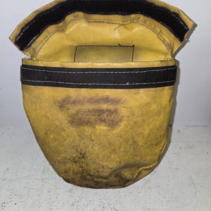 Op de afbeelding: Een gele tas met een zwarte klittenbandsluiting en een zwarte band met witte stiksels. De tas heeft donkere vlekken en lijkt gemaakt te zijn van een duurzaam, mogelijk waterdicht materiaal. De bovenklep is open.