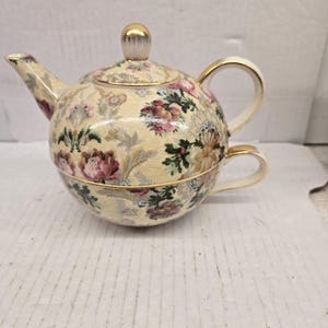 May include: A cream-coloured teapot and teacup set with a floral pattern. The teapot has a round shape, a lid, and gold-coloured handle and spout. The teacup fits around the teapot. The set features pink, purple, and green floral designs.
