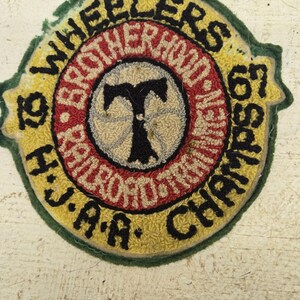Peut inclure: Un écusson vintage circulaire avec le texte "WHEELERS BROTHERHOOD 67 CHAMPIONS J.A.A." en lettres noires. L'écusson a un fond jaune avec un cercle intérieur rouge et blanc et une bordure verte. Le centre présente un "T" noir.