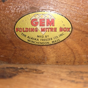 May include: An oval sticker on a wooden surface. The sticker is yellow with red text that reads "GEM FOLDING MITRE BOX." Below the text is "MFD. BY THE ALASKA FREEZER CO. INC. WINCHENDON, MASS."