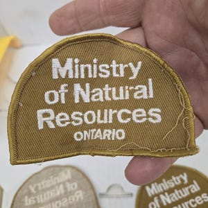 Peut inclure: Plusieurs écussons semi-circulaires beiges avec du texte brodé blanc. Les écussons indiquent « Ministry of Natural Resources ONTARIO ». Les écussons sont tenus dans une main sur un fond blanc.