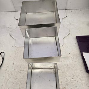 Puede incluir: Tres moldes cuadrados para hornear de acero inoxidable de diferentes tamaños están apilados. Los moldes tienen un diseño simple y utilitario con lados rectos y un acabado metálico brillante. Están dispuestos sobre una superficie clara.