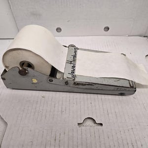 May include: A vintage grey metal paper roll dispenser featuring a horizontal bar with the handwritten text Wawa-Hardware. The device holds a large roll of white paper that feeds through a metal guide, showing signs of wear and aged paint.