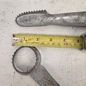 Peut inclure: Deux outils métalliques vintage avec des surfaces texturées. L'un des outils a un bord dentelé et mesure environ 13 cm de long. L'autre outil a une extrémité circulaire avec un bord dentelé. Un mètre ruban jaune est également visible.