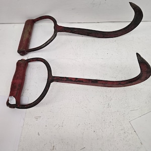 May include: Two antique red and black metal hay knives. Each tool features a curved blade and a D-shaped handle with a wooden grip. The blades are curved and the handles are painted red.