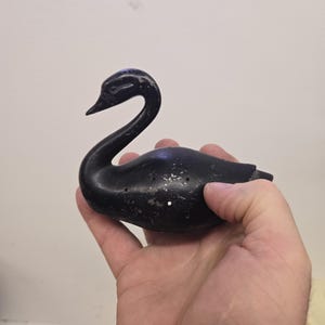 Puede incluir: Figura de cisne negro con cuello curvo y pico detallado. La figura tiene varios agujeros pequeños y parece estar hecha de un material oscuro. El cisne se sostiene en una mano sobre un fondo liso.