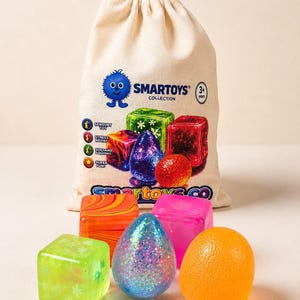 May include: A beige drawstring fabric bag printed with the text SMARTOYS COLLECTION and graphics of various sensory toys, accompanied by five loose sensory toys: a green cube with flower patterns, an orange and red marbled cube, a blue glittery egg, a pink cube, and an orange sphere.
