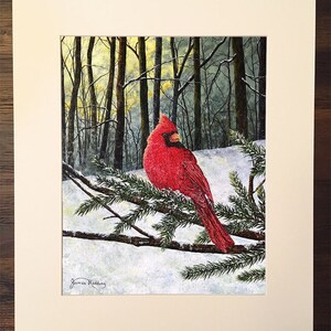 May include: A red cardinal perched on a snow-covered evergreen branch in a snowy forest. The cardinal is facing left and has a black eye and beak. The background is a snowy forest with bare trees. The artist's signature, "James Redding", is in the bottom left corner.