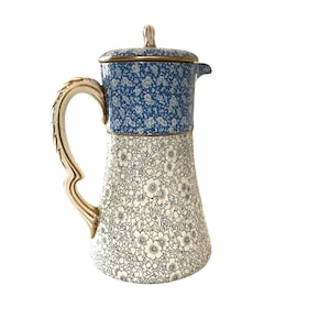May include: A white ceramic pitcher with a blue and white floral pattern. The pitcher has a gold handle and a lid.