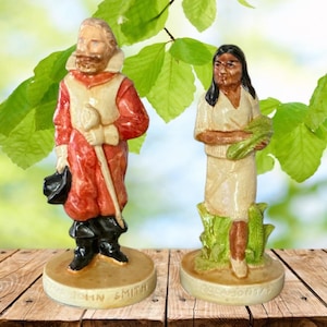 Può includere: Due figurine in ceramica, una raffigura John Smith in un costume rosso e bianco che tiene uno scettro e un cappello, l'altra raffigura Pocahontas in un abito bianco che tiene un mazzo di foglie verdi. Entrambe le figurine sono in piedi su una superficie di legno marrone.