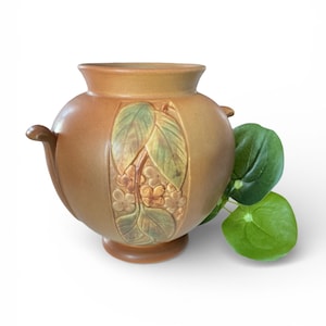 May include: A brown ceramic vase with a floral and leaf relief design. The vase has two small handles and a round base. Green leaves are next to the vase. The vase is a decorative item.