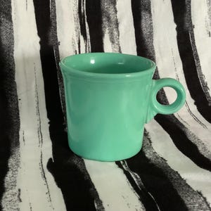 May include: A turquoise blue ceramic mug with a single handle. The mug has a rounded shape and a smooth, glossy finish.