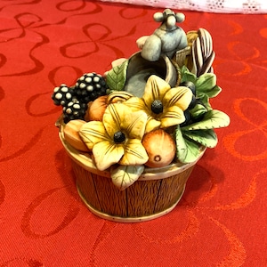 May include: A ceramic basket ornament filled with sculpted fruits and flowers. The basket has a brown wood-grain effect, and the arrangement includes yellow flowers, grapes, and peaches. A small mouse figurine sits on the edge.
