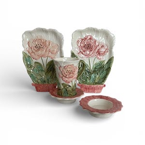 May include: A ceramic tea set featuring a floral design. The set includes two plates, a teacup, a saucer, and a small bowl. The plates and teacup have a raised peony design in pink, with green leaves and a red basket-weave base.
