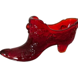 May include: A decorative glass shoe in a deep red hue. The shoe features a high heel and a pointed toe, with a floral pattern embossed on its surface. This ornamental piece is designed to resemble a shoe.