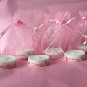 K&ouml;nnte beinhalten: F&uuml;nf wei&szlig;e Teelichter mit rosa Beschriftung. Die Kerzen befinden sich in rosa Organzas&auml;ckchen. Der Text auf den Kerzen lautet "Jackie's Hen Party 26.03.2016".