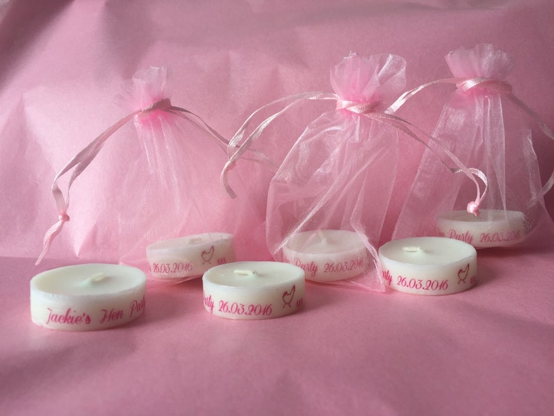 K&ouml;nnte beinhalten: F&uuml;nf wei&szlig;e Teelichter mit rosa Beschriftung. Die Kerzen befinden sich in rosa Organzas&auml;ckchen. Der Text auf den Kerzen lautet "Jackie's Hen Party 26.03.2016".