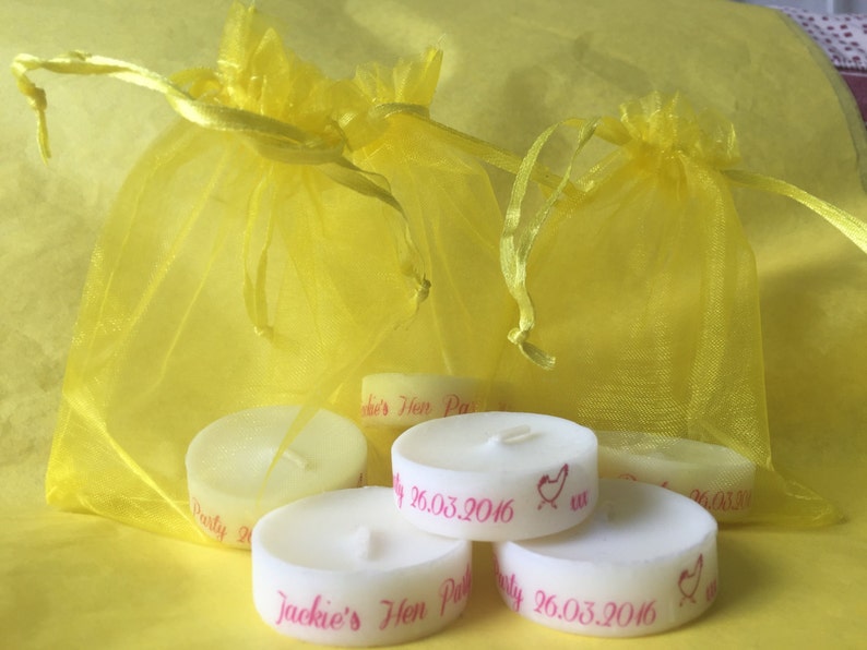 K&ouml;nnte beinhalten: Sechs wei&szlig;e Teelichter mit rosa Beschriftung. Die Kerzen befinden sich in gelben Organzas&auml;ckchen. Der Text auf den Kerzen lautet "Jackie's Hen Party 26.03.2016".