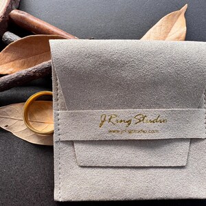 A gray fabric pouch with gold text and a website address, along with a pair of gold rings and some dried leaves, arranged on a dark surface.