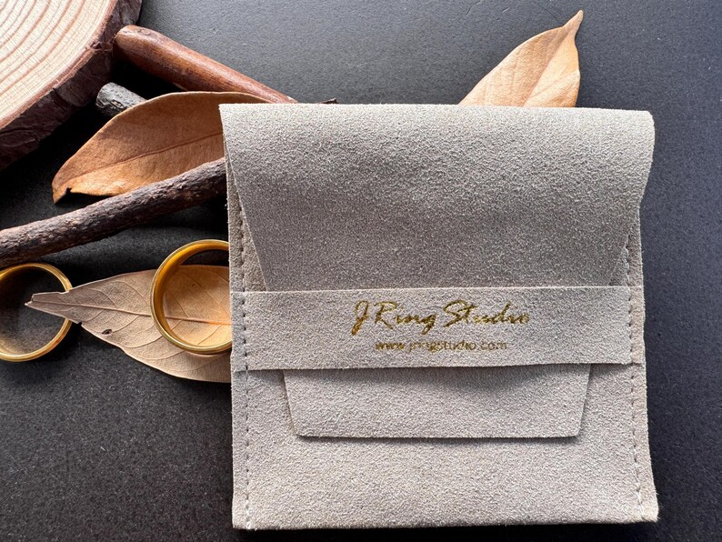 A gray fabric pouch with gold text and a website address, along with a pair of gold rings and some dried leaves, arranged on a dark surface.