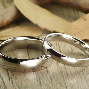 May include: Two polished silver wedding bands, one slightly larger than the other, rest on a wooden surface with dried leaves. The rings reflect light, showcasing their smooth, rounded design.