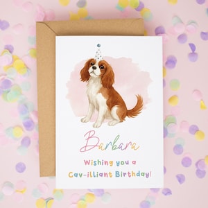 May include: A white greeting card featuring a watercolor illustration of a brown and white Cavalier King Charles Spaniel wearing a small party hat. The text reads Barbara, Wishing you a Cav-illiant Birthday! The card is paired with a brown envelope.