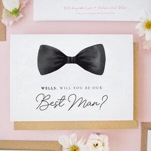 May include: A white greeting card featuring a black watercolor bow tie illustration centered at the top. Below the bow tie, the text reads: WELLS, WILL YOU BE OUR Best Man? The card is presented against a light pink background with white flowers.
