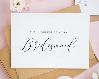 May include: A white rectangular card with black text that reads THANK YOU FOR BEING MY Bridesmaid. The card is placed on top of a brown cardboard envelope. The design features a clean, minimalist aesthetic with a simple serif and script font.