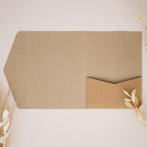 May include: A brown paper pocket folder with a smaller pocket on the right. The folder is open, showing the inside. Dried plants are placed on the left and right, against a light pink background.